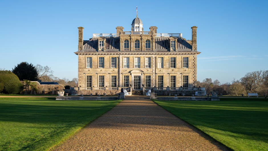 The image depicts the front view of a grand, historic stone mansion with multiple large windows and a central entrance, situated at the end of a long, straight gravel pathway. The building features chimney stacks, a dome-shaped central roof structure, and a balustrade along the top level, with a clear blue sky overhead. The surrounding landscape includes well-maintained green lawns, a few trees in the background, and benches on either side of the pathway. This setting suggests a picturesque environment suitable for professional house removals or home relocation projects. Occasionally, furniture, packing materials, or moving equipment operated by Man with Van Addington may be involved in the process of furniture transport and packing within or around such a property during a local move or loading process.