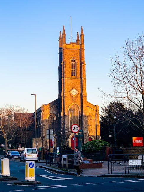 A tall, historic stone church with a prominent clock face and narrow arched windows, situated on a street corner during late afternoon with warm sunlight casting an orange hue on the building. The church features a rectangular tower topped with pinnacles and a flagpole, surrounded by leafless trees and street furniture including road signs, bollards, and traffic barriers. In the foreground, a pedestrian carrying a bag walks along the paved sidewalk, while cars wait at a nearby intersection. The scene captures a typical urban setting aligned with house removals and home relocation logistics, demonstrating the church's position as a local landmark for several property moving services in the Addington area, as promoted by Man with Van Addington. The setting underscores the importance of careful planning during furniture transport and packing as part of professional removals.