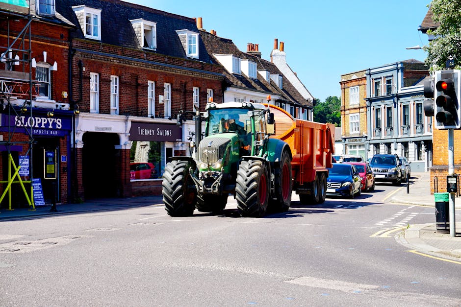A large green tractor with an orange waste collection trailer attached is positioned at an intersection within a residential area during daytime. The tractor is on the road, partially blocking the crosswalk, with its front facing towards the viewer. The surrounding environment includes brick buildings with retail shops on the ground floor, such as a sports bar and a hair salon, as well as parked cars along the side of the street. Bright sunlight illuminates the scene, casting shadows on the pavement. A traffic light displays a red signal at the corner, and a green trash bin is visible on the sidewalk. The scene captures a typical urban setting where heavy machinery might be used for moving or waste disposal, relevant to house removals, packing, or disposal services offered by Man with Van Addington.