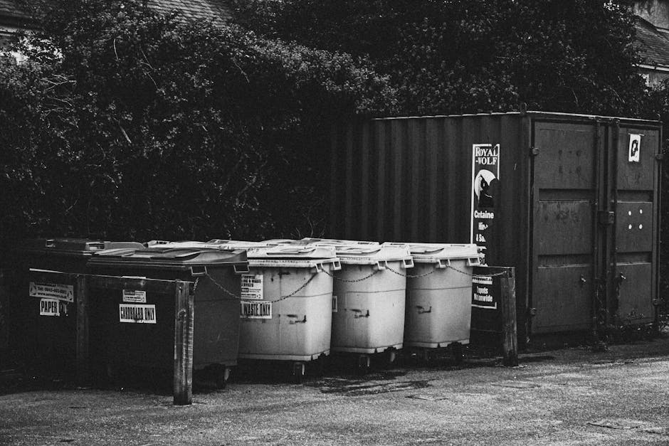 A row of four large white wheelie bins with closed lids, positioned next to a wooden post on a paved surface, with a second row of black waste containers behind them. The white bins are labeled for waste disposal, and they are placed between a dark large skip container and a black metal fence. The background includes dense foliage and trees, partially visible behind the skip. The scene appears to be outside a residential property, with the bins likely used for household waste collection during a house removal or relocation. The setting is in natural daylight, and the image remains in black and white, emphasizing the utilitarian aspect of waste management in the context of moving or packing processes, as potentially handled by Man with Van Addington during a home relocation.