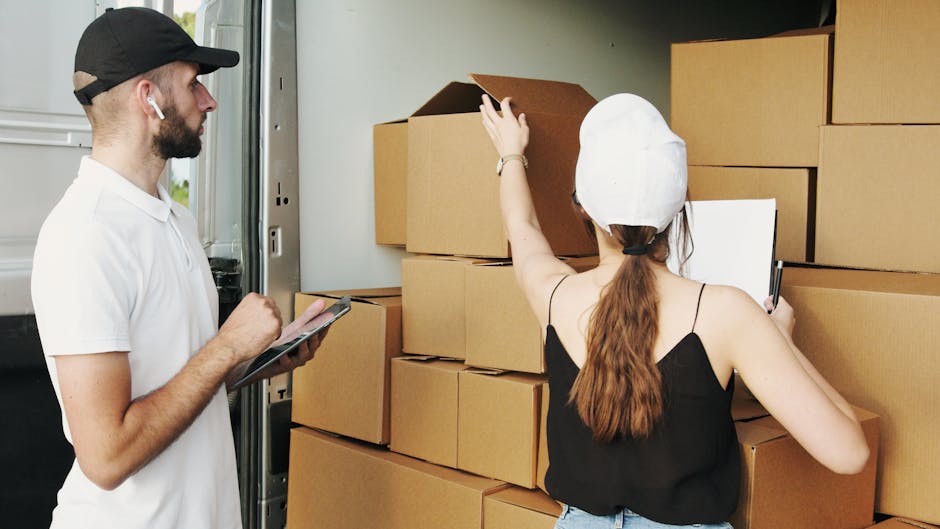 A man wearing a black cap and white t-shirt stands next to an open delivery van, holding a clipboard and pen, while a woman with long brown hair, dressed in a black sleeveless top and white cap, is inside the van organizing and inspecting packed cardboard boxes, some of which are stacked inside the van and others on a pavement nearby. The woman is pointing towards a box on the stack, possibly planning the loading process during a home relocation. The van's interior is partially visible, showing additional boxes and packing materials, with natural daylight illuminating the scene. This image depicts the packing and loading stage of furniture transport and house removals, illustrating professional moving services, as seen by the involvement of [COMPANY_NAME] in managing the logistics of a house removal.