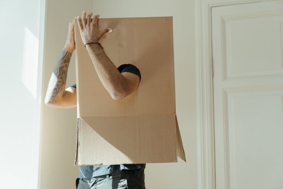 A person wearing a black short-sleeved shirt and grey pants is carrying a large cardboard box with a circular hole cut out on the side, likely for a house move or packing process. The individual has a tattooed arm visible, and their face is obscured by the box. The box appears to be a standard moving carton, suitable for packing household items, and is held securely with both hands, one at the top and the other supporting the side. The scene takes place indoors, next to a white door and a cream-colored wall, with natural light illuminating the space. Nearby, additional packing materials such as cardboard boxes and plastic wraps might be on the floor or nearby surfaces, indicating a home relocation environment. The setting suggests active furniture transport or packing, typical of professional removals services provided by companies like Man with Van Addington, as they assist with efficient and careful home moves, including loading and transport in moving vans.