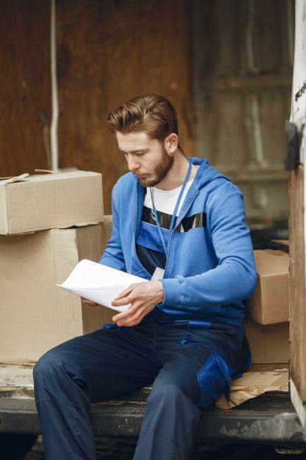 A man with dark hair, wearing a black t-shirt, stands inside a room near an open window with wooden frames, looking outside. Visible on the bed are several packed cardboard boxes, some with packing ta