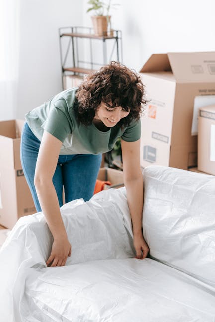 A woman with curly brown hair wearing a green t-shirt and blue jeans is standing inside a room, actively wrapping or preparing a large piece of furniture, possibly a sofa, with white protective covering during a home relocation. Behind her, there are cardboard boxes, some open, and a metal and wooden shelving unit holding a potted plant and small items. The room has natural lighting from an unseen window, and the floor is wooden. Visible in the background are unpacked boxes and additional packing materials, indicating an ongoing packing or moving process. This scene reflects the household packing phase typical of furniture transport and house removals, with the woman managing the preparation of items as part of a professional moving or packing service, such as offered by Man with Van Addington.