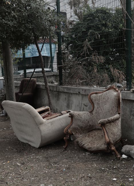 Two antique upholstered armchairs with wooden frames and fabric upholstery, positioned outdoors on a gravel surface, leaning against a concrete wall and a metal fence. One chair appears more frayed and worn, while the other has a slightly cleaner appearance. Behind the chairs, there are trees and bushes with dense foliage, and a glimpse of a building with windows and a blue exterior. The chairs are part of an outdoor storage area or discarded furniture collection, reflecting the process of home relocation or furniture removal. The scene is lit by natural daylight, with shadows cast by nearby objects.