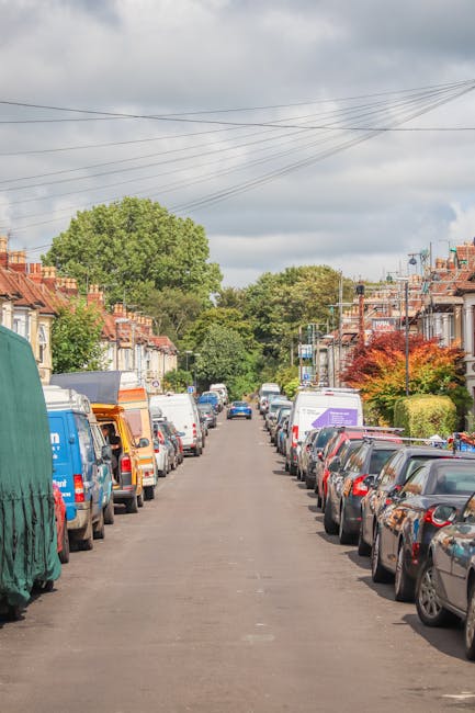 A narrow residential street in Addington lined with parked cars on both sides, including a mix of small hatchbacks, larger vans, and delivery vehicles. The street is paved with asphalt and features terraced houses with brick facades and tiled roofs, some with small front gardens or hedges. Several houses have visible bay windows, and a few trees and shrubs are present along the pavement, with one tree displaying vibrant red foliage. Overhead, multiple electrical wires run parallel to the street, connected to utility poles. The sky is overcast with grey clouds, suggesting typical urban housing and parking conditions suitable for local house removals, such as those provided by Man with Van Addington, experienced in logistics and furniture transportation during home relocations.