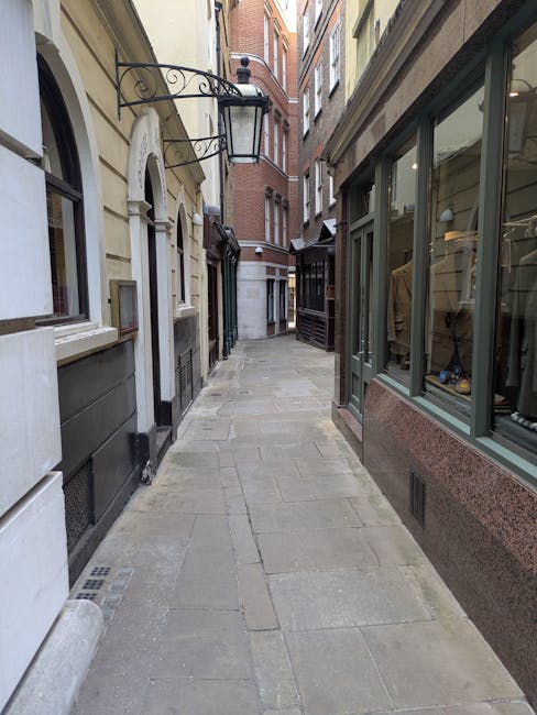 A narrow European street with paving stones, surrounded by multi-story buildings with brick and stone facades, some with large window displays and decorative wrought iron features. On the right, a shop window displays clothing and accessories, while on the left, residential doors and windows are visible. The street is mostly empty, with no pedestrians or vehicles visible, showing a typical setting for home relocation or moving services in Addington as provided by Man with Van Addington. The scene captures the outdoor environment where furniture transport and packing might take place during a house removal, with natural lighting highlighting the urban architecture.