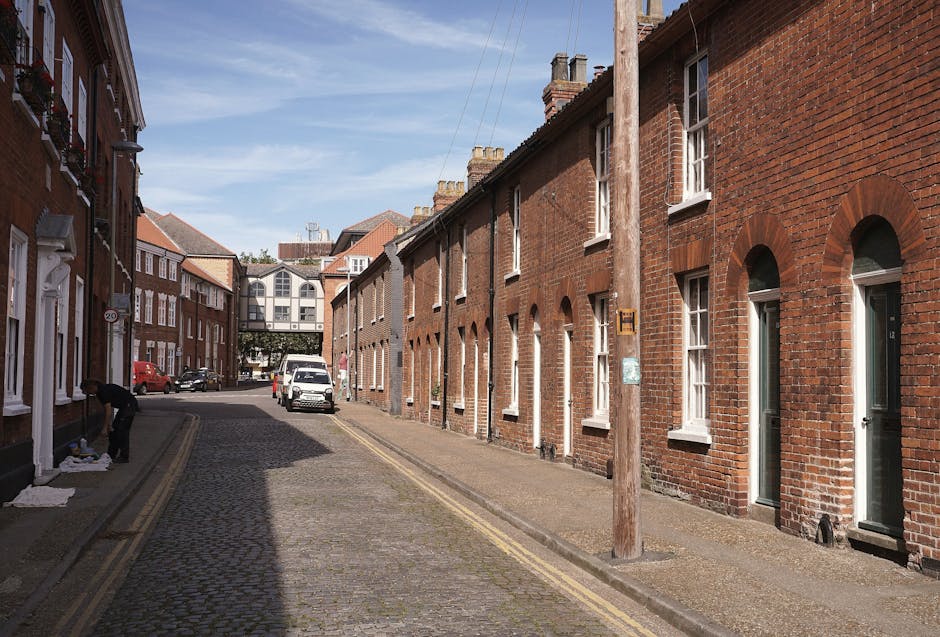 A narrow residential street in Addington lined with parked cars on both sides, including a mix of small hatchbacks, larger vans, and delivery vehicles. The street is paved with asphalt and features terraced houses with brick facades and tiled roofs, some with small front gardens or hedges. Several houses have visible bay windows, and a few trees and shrubs are present along the pavement, with one tree displaying vibrant red foliage. Overhead, multiple electrical wires run parallel to the street, connected to utility poles. The sky is overcast with grey clouds, suggesting typical urban housing and parking conditions suitable for local house removals, such as those provided by Man with Van Addington, experienced in logistics and furniture transportation during home relocations.