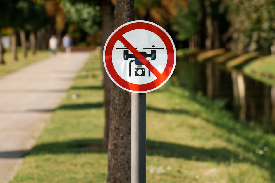 A circular traffic sign with a white background, a red border, and a black drone icon crossed out by a red diagonal line, indicating no drone flying allowed. The sign is mounted on a grey metal pole s