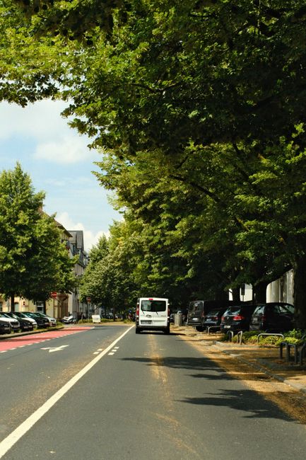 Image of a busy town centre street during daytime, featuring a row of multi-story commercial buildings with brick and plaster facades, some with shopfronts such as a customer service point and a pharm