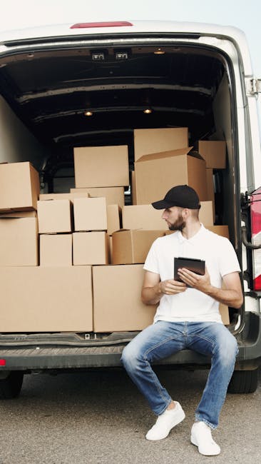 A man wearing a baseball cap, black gloves, and a black padded vest over a blue long-sleeve shirt stands outdoors next to an open white moving van, which has a brown wooden panel on its side. He is po