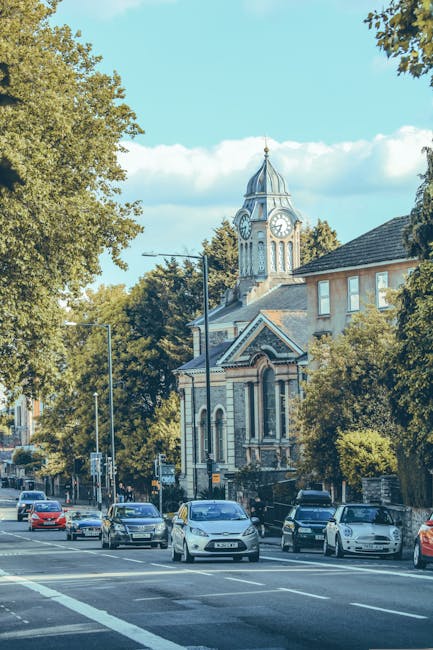 A street scene in Addington displaying a row of parked cars along the pavement, with a large historic building featuring a prominent clock tower with a spire visible in the background, surrounded by mature trees with green foliage. In the foreground, a moving van from Man with Van Addington is parked on the side of the road, with its sliding door open and several cardboard boxes, wrapped furniture pieces, and packing materials ready for loading. The areas around the van are set up for a home relocation, with items strategically placed to facilitate efficient furniture transport and packing during the moving process. The scene is bathed in natural daylight, showing clear skies with a few clouds, emphasizing the residential and community atmosphere typical for a house removal in the Addington area.