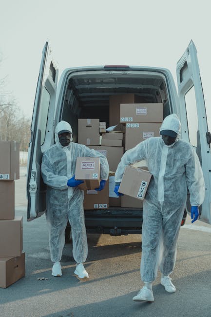Two movers from Man with Van Addington, dressed in white protective suits, gloves, and face coverings, are unloading cardboard boxes from the back of a white moving van parked outdoors on a paved surface. The van’s rear doors are open, revealing neatly stacked boxes of various sizes, some labeled with shipping information. One mover, on the left, is holding a small cardboard box, while the other, on the right, is carrying a larger box. Several additional boxes are on the ground nearby, ready for transport into a property. The scene takes place during daytime under clear lighting conditions, with a blurred background that suggests an urban or suburban environment. This image depicts the process of home relocation, part of a professional removals service, involving packing, loading, and furniture transport with the use of moving equipment like robust boxes and appropriate safety gear, emphasizing the logistics of quick and efficient relocation services offered by Man with Van Addington for emergency clearances or scheduled moves.