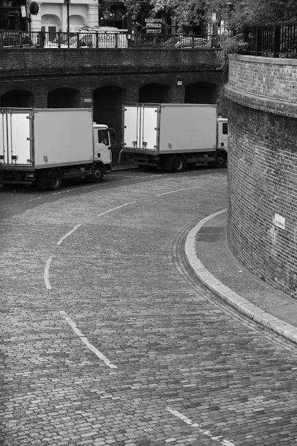 A black-and-white photograph of a loading area featuring two white box trucks parked parallel to a brick wall, with the rear ends facing a curved cobblestone driveway. The driveway has white dashed lines indicating parking guidance, and the trucks are positioned close to a low bridge or archway beneath a residential or commercial building with a balcony and railing. The scene is outdoors with natural lighting, and the trucks are part of a home relocation or moving process. Visible on the right side are the brick wall and a small sign, suggesting a designated loading zone. The image reflects a typical setting for furniture transport or packing and moving activities, with the trucks prepared for loadings associated with house removals, supported by the presence of equipment such as straps or blankets that may be nearby, indicative of careful handling during a move. Man with Van Addington handles these relocation tasks, ensuring efficient and secure transport of household items.