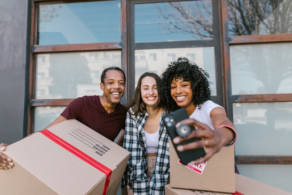 Three young adults standing outside a property with large glass windows, smiling and taking a selfie together. The person on the left is holding a cardboard box sealed with red tape labeled 'moving boxes,' while the person on the right is holding a similar box and a smartphone in an extended arm. The individual in the middle wears a plaid shirt and has long brown hair. The background shows the exterior of a building with a cityscape reflected in the windows. The scene depicts a casual moment during a house removal or packing process, likely part of a student move-out arrangement as associated with Man with Van Addington, focusing on friendly teamwork in furniture transport and moving logistics.